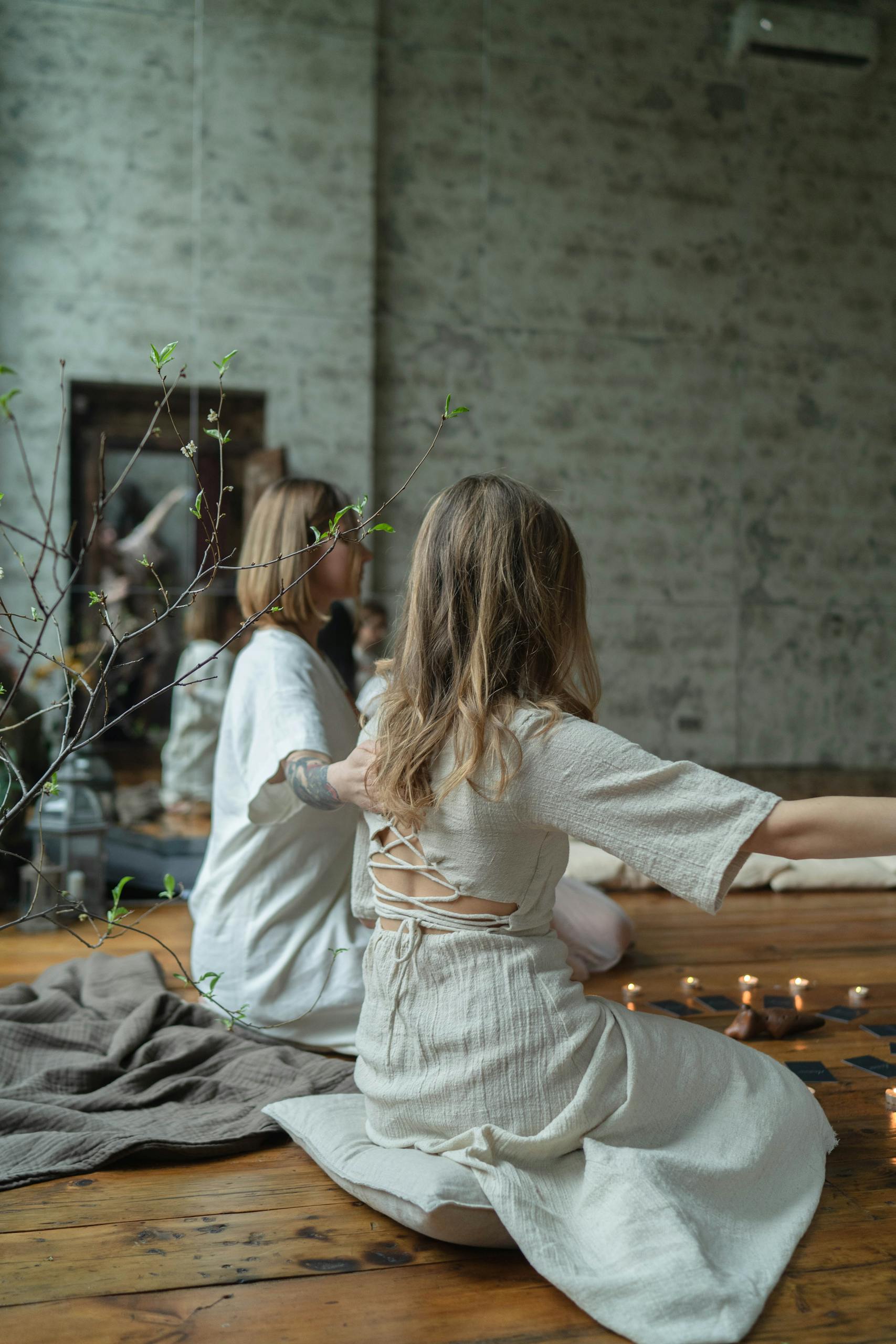 Two women in white dresses meditating indoors with candles and serene ambiance.